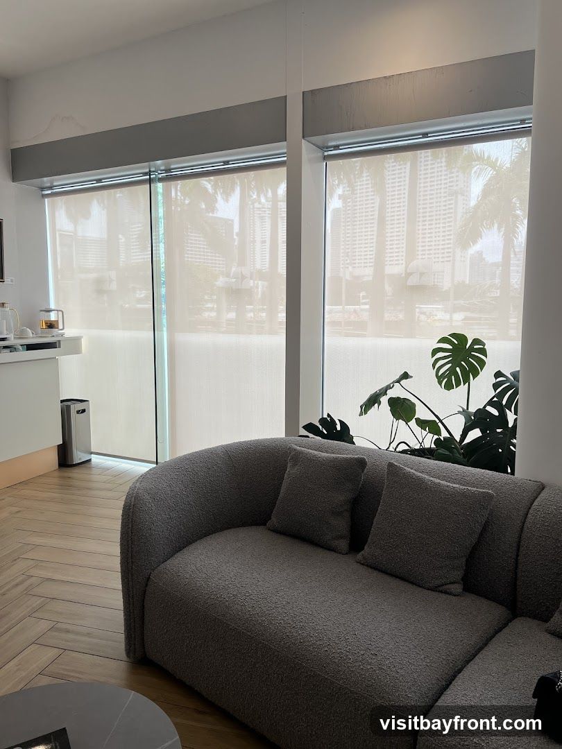 A comfortable grey textured sofa with two pillows sits in a modern waiting area. Large windows with sheer blinds reveal a cityscape with palm trees. A large monstera plant adds a touch of greenery. To the left, a white counter has a kettle and some cups, and a silver trash can is visible. The floor has a herringbone wood pattern.