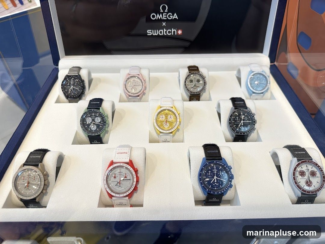 A display case featuring multiple Omega x Swatch watches. The watches are arranged in three rows, with each watch presented on a white stand. The top row shows four watches in black, pink, silver, and light blue. The middle row displays watches in green, yellow, and dark gray. The bottom row features watches in beige, red, dark blue, and white with red accents. The top of the display has a black background with the Omega and Swatch logos and the text "OMEGA x swatch+".