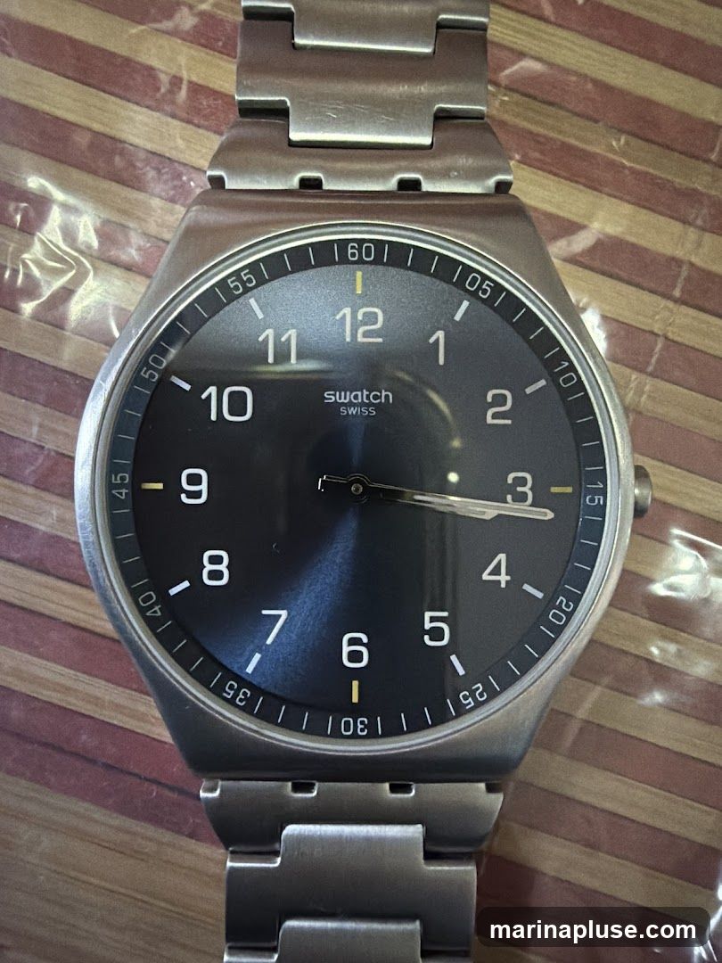 A close-up, top-down view of a Swatch watch with a dark gray face and a silver metal band. The watch has white Arabic numerals for the hours and a minute track around the outer edge. The brand name "swatch SWISS" is visible on the face. The watch is lying on a surface with a striped pattern.