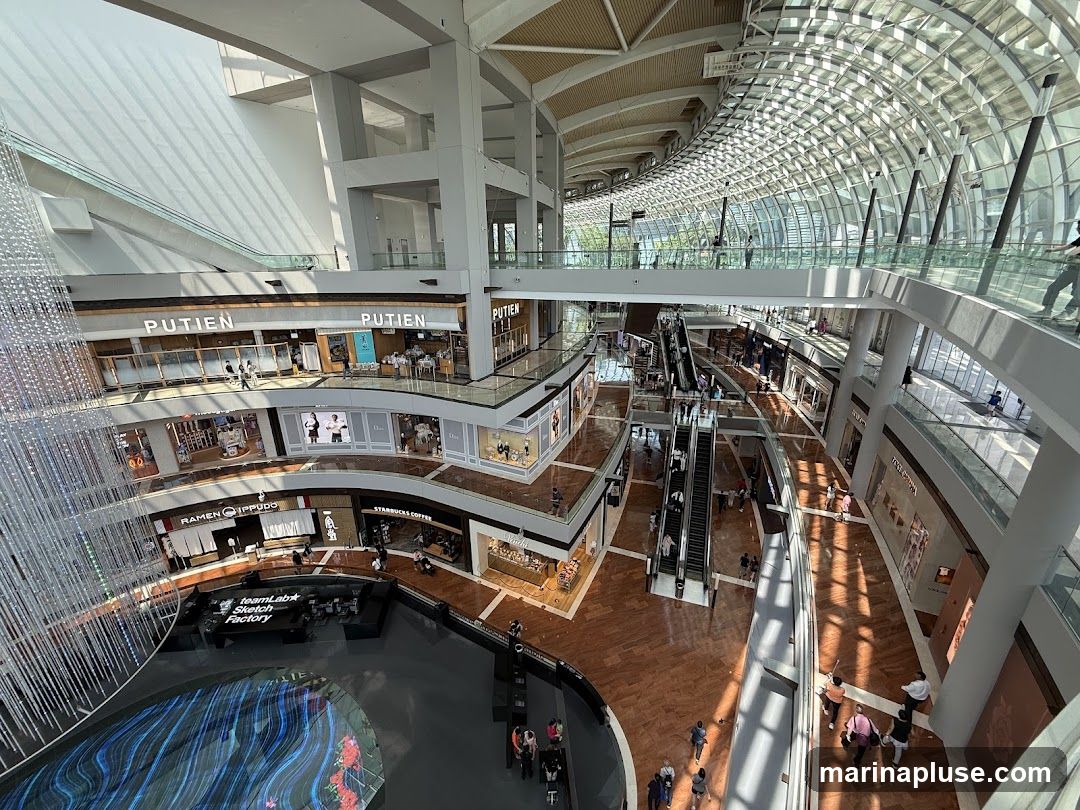 An aerial view of the interior of a multi-level shopping mall. The mall features a large, open atrium with escalators and walkways connecting different floors. Sunlight streams in through a curved glass ceiling, casting shadows on the wooden floors. Various shops are visible on the different levels, including 'PUTIEN', 'RAMEN IPPUDO', and 'STARBUCKS COFFEE'. A large digital display or art installation is present in the lower section of the atrium.