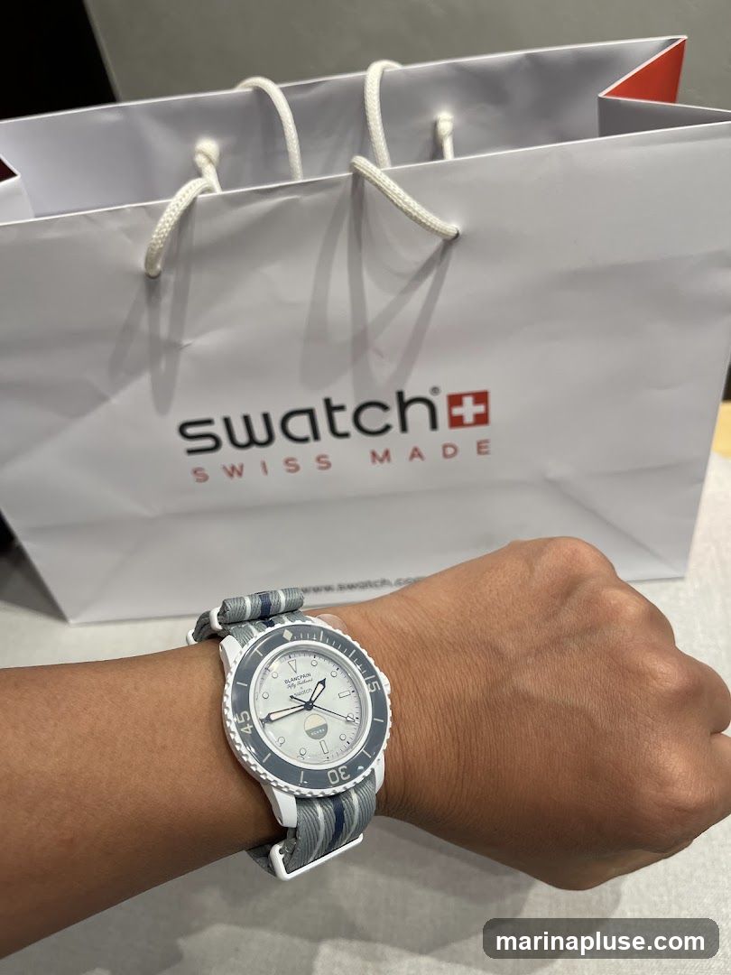 A person's wrist wearing a white and grey striped watch with a white watch face. In the background, a white shopping bag with the Swatch logo and "SWISS MADE" text is visible.