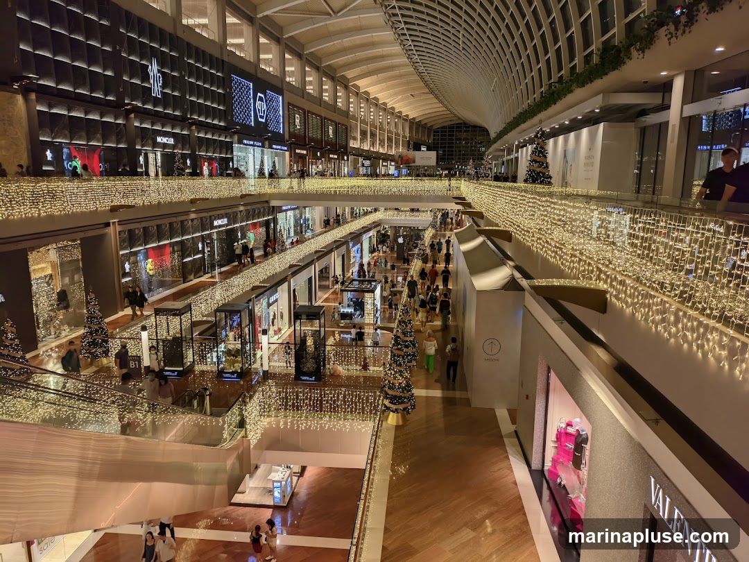 The image shows the interior of a multi-level shopping mall, adorned with numerous strings of warm white fairy lights along the railings and balconies. Several Christmas trees are visible, some decorated and placed throughout the mall. The shops on each level have illuminated displays and signage, including brands like Moncler, Philipp Plein, and Guess. People are seen walking around and browsing the shops. The architecture features a high, curved ceiling and wooden flooring.