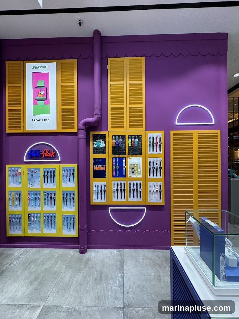 A vibrant purple wall serves as a backdrop for a Swatch store display. Yellow shutters are mounted on the wall, and a purple pipe runs vertically. Several display cases showcase a variety of Swatch watches. One large poster features a Swatch watch with the text "BREAK FREE!". Neon lights in the shape of a semicircle are also visible. A glass display case in the foreground contains watch boxes.
