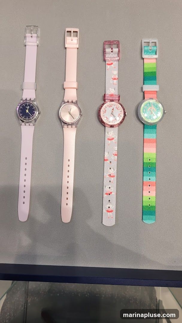 Four watches are displayed on a gray surface. From left to right, the first watch has a light purple band and a dark blue face. The second watch has a light pink band and a rose gold face. The third watch has a light gray band with a pattern of ballerinas and a pink glittery bezel. The fourth watch has a colorful striped band with green, pink, and blue stripes and a light green face.