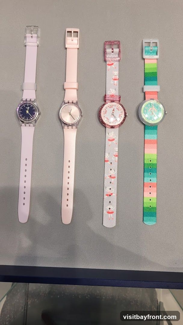Four watches are displayed on a gray surface. From left to right, the first watch has a light purple band and a dark blue face. The second watch has a light pink band and a rose gold face. The third watch has a light gray band with a pattern of ballerinas and a pink glittery bezel. The fourth watch has a colorful striped band with green, pink, and blue stripes and a light green face.