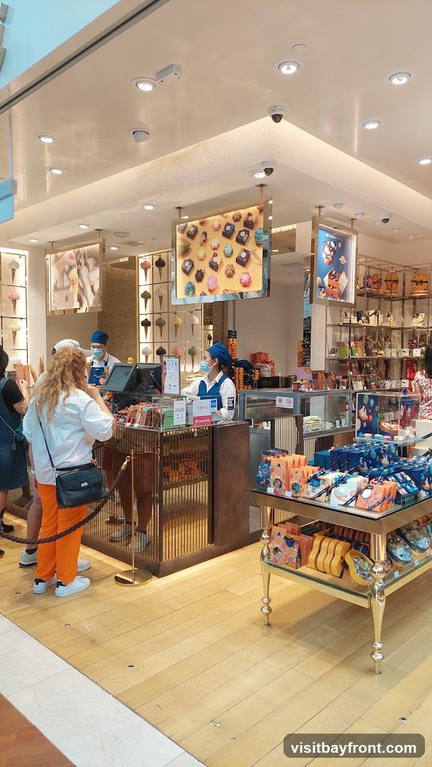 An interior view of a brightly lit shop selling ice cream and chocolates. Customers are being served by staff behind a counter. Display shelves are filled with packaged chocolates and other sweets. Pictures of ice cream cones and chocolates are displayed on the walls and hanging from the ceiling.