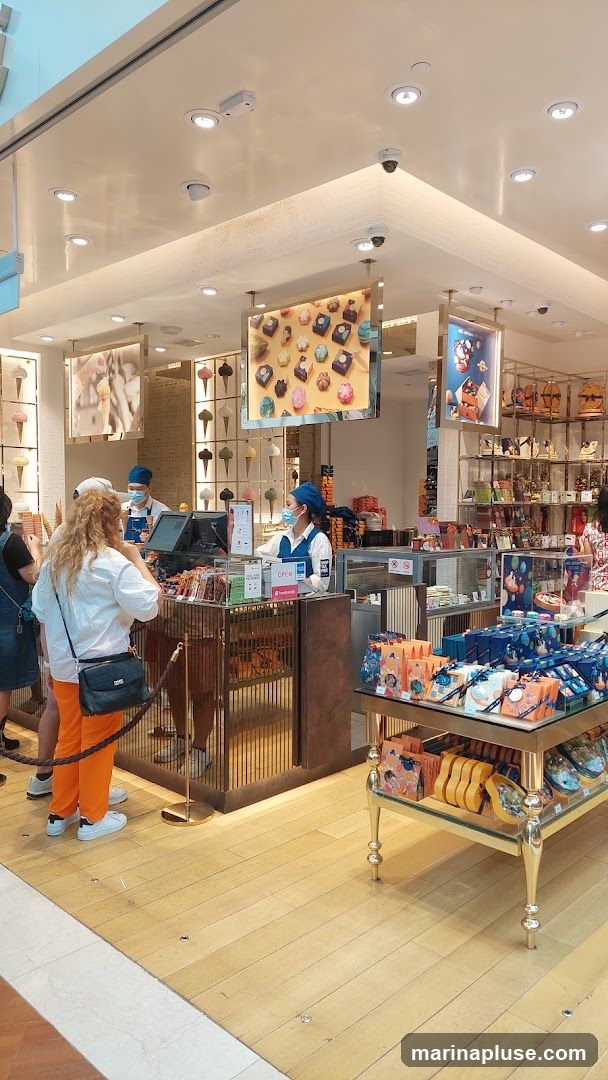 An interior view of a brightly lit shop selling ice cream and chocolates. Customers are being served by staff behind a counter. Display shelves are filled with packaged chocolates and other sweets. Pictures of ice cream cones and chocolates are displayed on the walls and hanging from the ceiling.