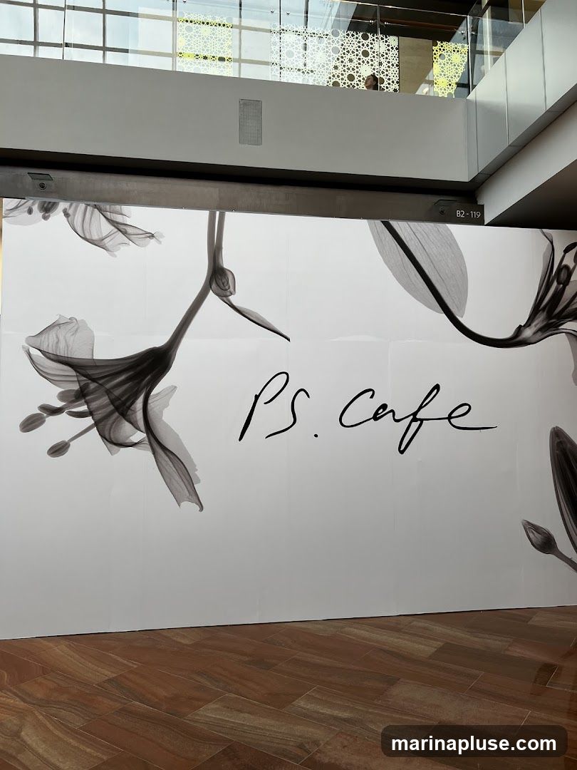 A large white mural with a black floral design and the text "PS. Cafe" in cursive handwriting. The mural is displayed in an indoor setting with a wooden floor and a balcony with decorative screens in the background.