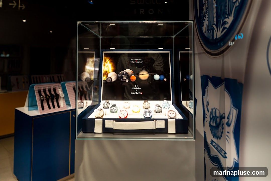 A display case showcasing Omega x Swatch watches. The background features a graphic of planets and the sun, with text reading "BIOCERAMIC MOONSWATCH COLLECTION". Below, a collection of watches are arranged in a blue velvet-lined case. To the left, another display shows several watches on stands.