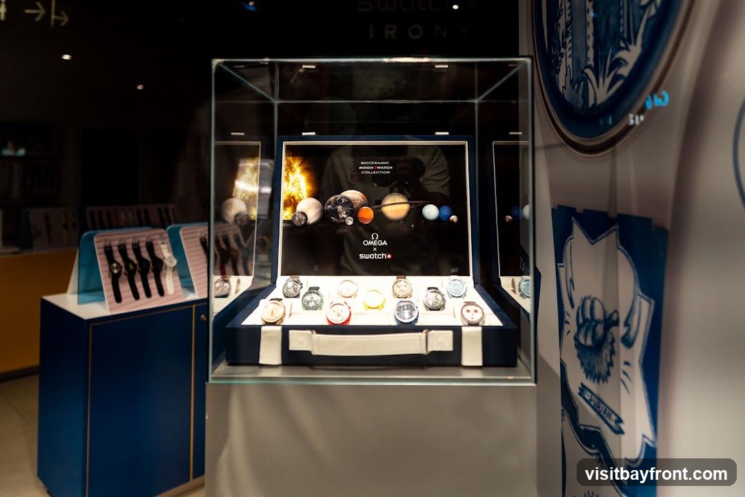 A display case showcasing Omega x Swatch watches. The background features a graphic of planets and the sun, with text reading "BIOCERAMIC MOONSWATCH COLLECTION". Below, a collection of watches are arranged in a blue velvet-lined case. To the left, another display shows several watches on stands.