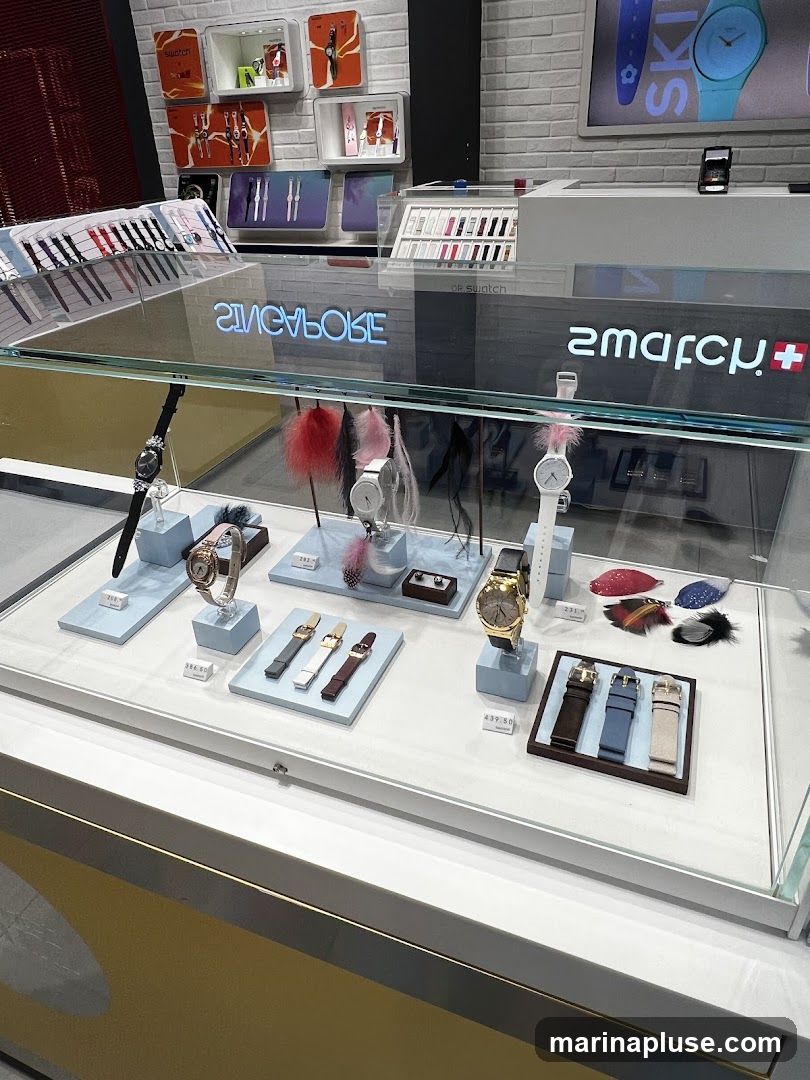 A display case filled with various Swatch watches and accessories. The watches are presented on light blue stands, with some featuring decorative feathers. In the background, shelves display more watches and branded promotional materials. A neon sign reflecting in the glass reads "SINGAPORE" and "SWATCH".