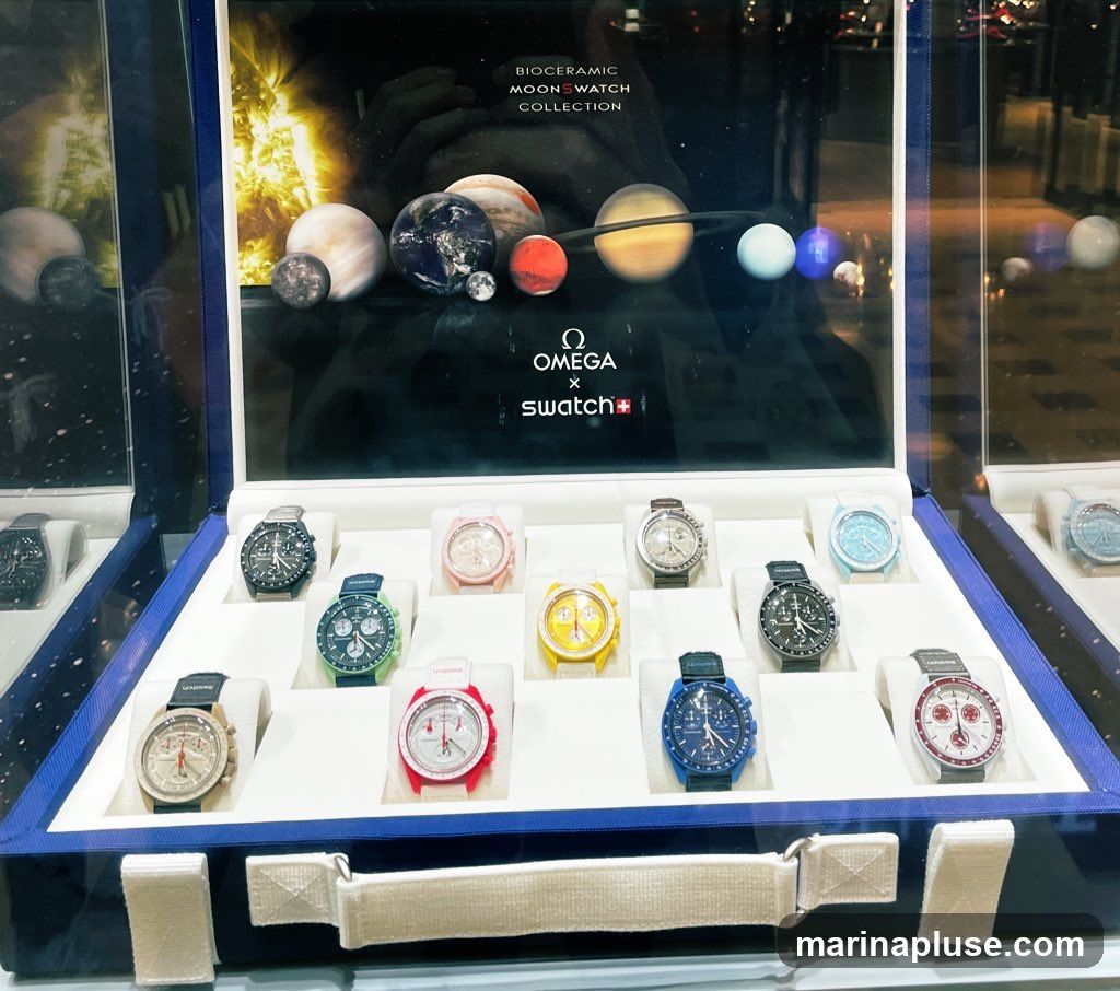 A display case showcasing the Bioceramic MoonSwatch Collection by Omega x Swatch. The collection features watches inspired by planets, with various colors and designs. The backdrop features images of planets and the Omega and Swatch logos.