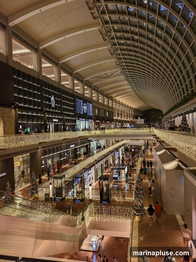 The image shows the interior of a multi-level shopping mall, adorned with numerous strings of warm white lights and decorated Christmas trees. The architecture features a high, arched ceiling with a grid-like structure and a large skylight. Shops with illuminated signage are visible on different levels, including Moncler and Dolce & Gabbana. People are seen browsing and walking through the mall.
