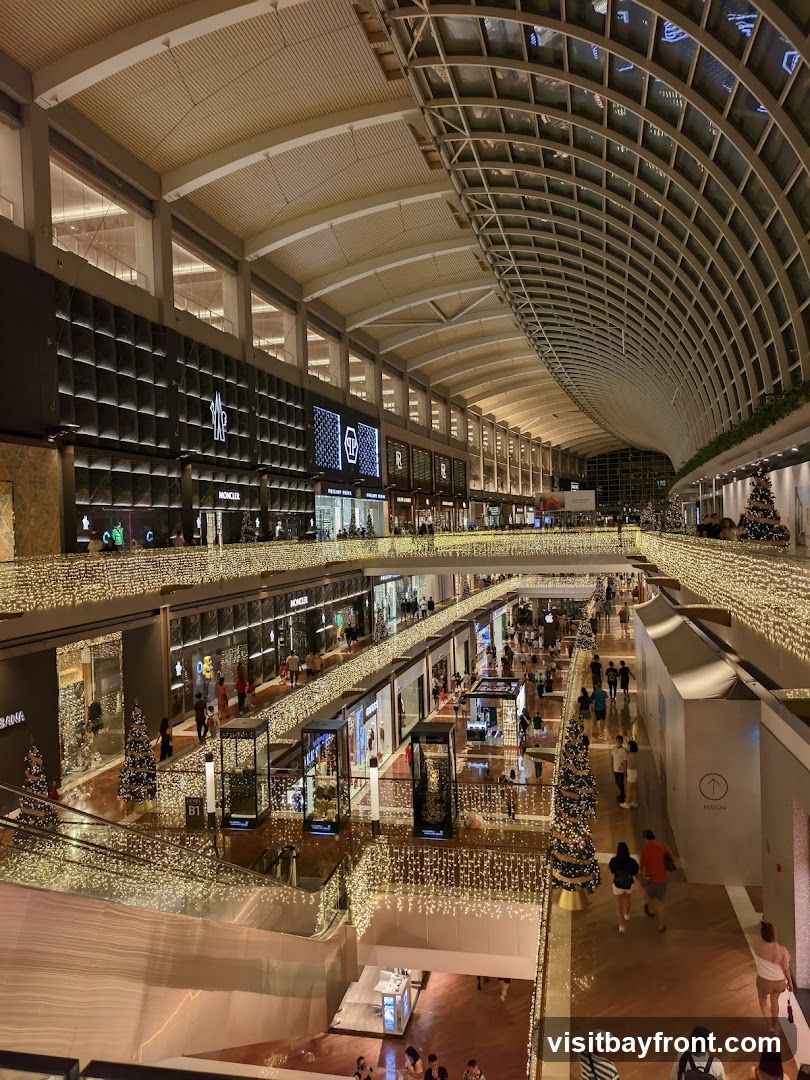 The image shows the interior of a multi-level shopping mall, adorned with numerous strings of warm white lights and decorated Christmas trees. The architecture features a high, arched ceiling with a grid-like structure and a large skylight. Shops with illuminated signage are visible on different levels, including Moncler and Dolce & Gabbana. People are seen browsing and walking through the mall.