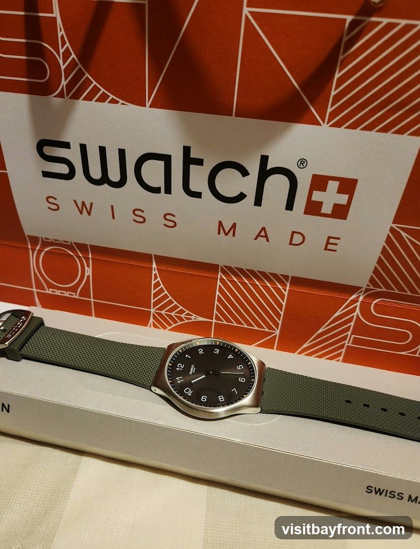 A Swatch watch with a dark face and green strap is displayed on a light-colored box. Behind the watch is a red and white Swatch shopping bag with the brand name and "SWISS MADE" printed on it.