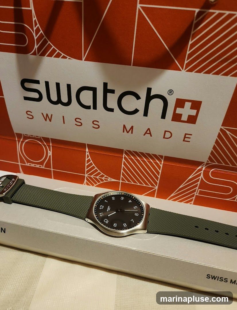 A Swatch watch with a dark face and green strap is displayed on a light-colored box. Behind the watch is a red and white Swatch shopping bag with the brand name and "SWISS MADE" printed on it.