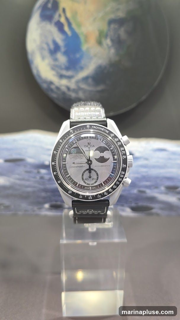 A close-up shot of an Omega Swatch MoonSwatch watch displayed on a clear stand. The watch has a silver dial with moon phase indicators and the words "Speedmaster" and "MOONSWATCH" visible. The background features a blurred image of Earth and a lunar landscape.