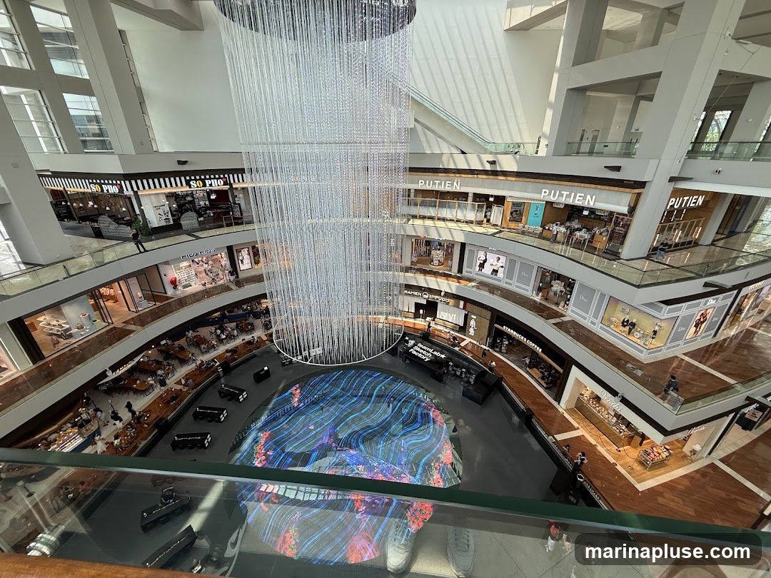 An interior view of a modern shopping mall, showcasing multiple levels of shops and walkways. A large, intricate chandelier hangs from the ceiling, and a vibrant digital display is visible in the center of the atrium. Shops like 'So Pho', 'Putien', 'Dior', and 'Starbucks Coffee' are identifiable. People are seen browsing and dining.