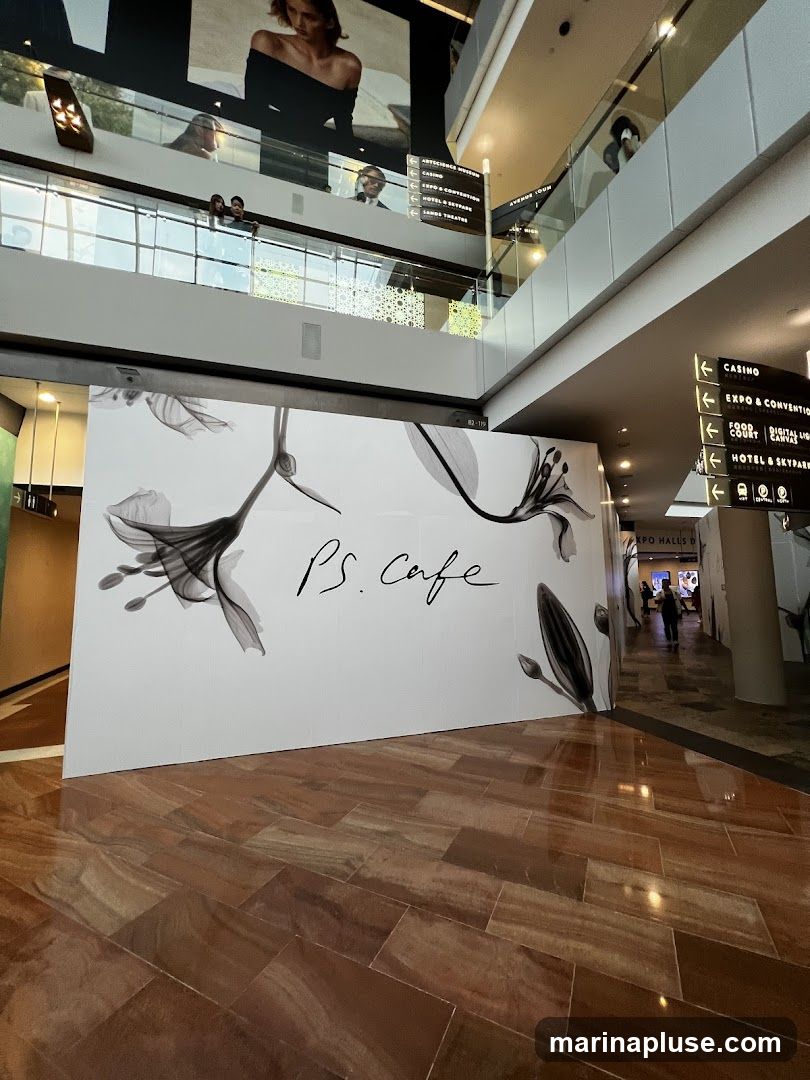 A large white panel with the text "PS. Cafe" in cursive script and floral illustrations is displayed in a modern indoor space with marble flooring. In the background, directional signs indicate various locations like "CASINO", "EXPO & CONVENTION", "FOOD COURT", and "HOTEL & SKYPARK".