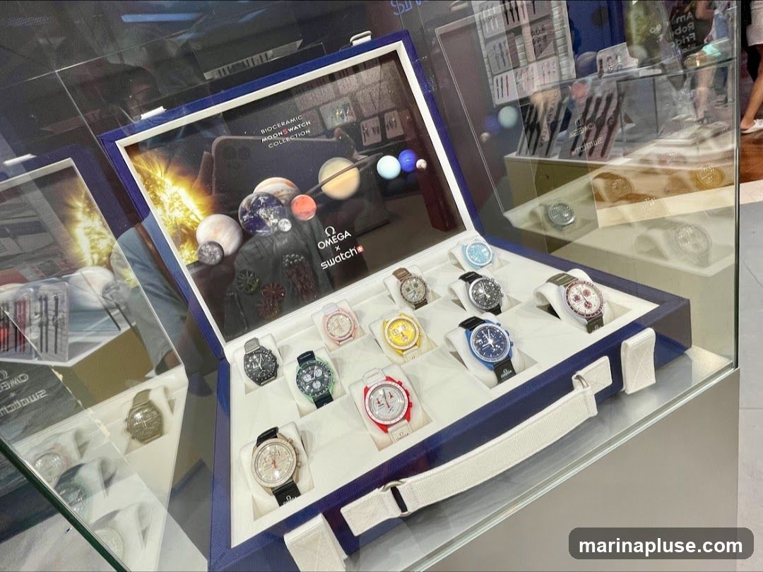 A display case showcasing the Omega x Swatch Bioceramic MoonSwatch watch collection. The case is open, revealing multiple watches arranged in compartments. The backdrop of the case features imagery of planets and the sun, with the Omega and Swatch logos prominently displayed. Several watches are visible, including models in black, green, blue, red, yellow, and white. Additional watch displays and products are visible in the background through reflections and adjacent cases.