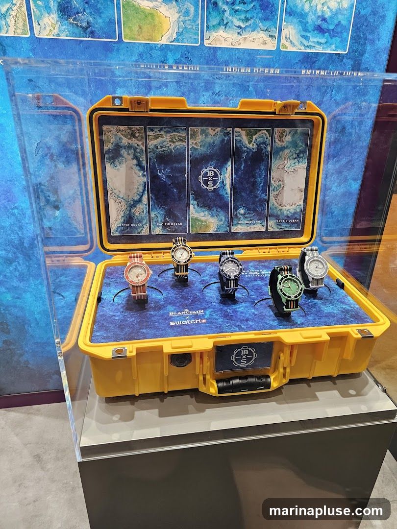 A yellow Pelican-style case is open, displaying five watches from the Swatch x Blancpain Ocean Collection. Each watch is placed on a blue mat featuring images of ocean depths. Above the watches, the lid of the case displays five panels, each depicting a different ocean with text labels such as "ARCTIC OCEAN", "PACIFIC OCEAN", "ATLANTIS", "INDIAN OCEAN", and "ANTARCTIC OCEAN". The Swatch and Blancpain logos are visible on the mat. The display is enclosed in a clear acrylic case, with a background featuring maps of various oceans.