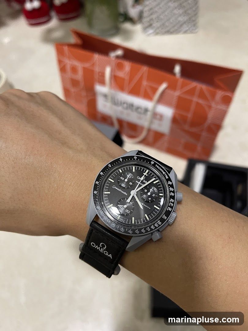A person's wrist wearing a grey and black watch with an Omega strap. In the background, an orange Swatch shopping bag is visible.