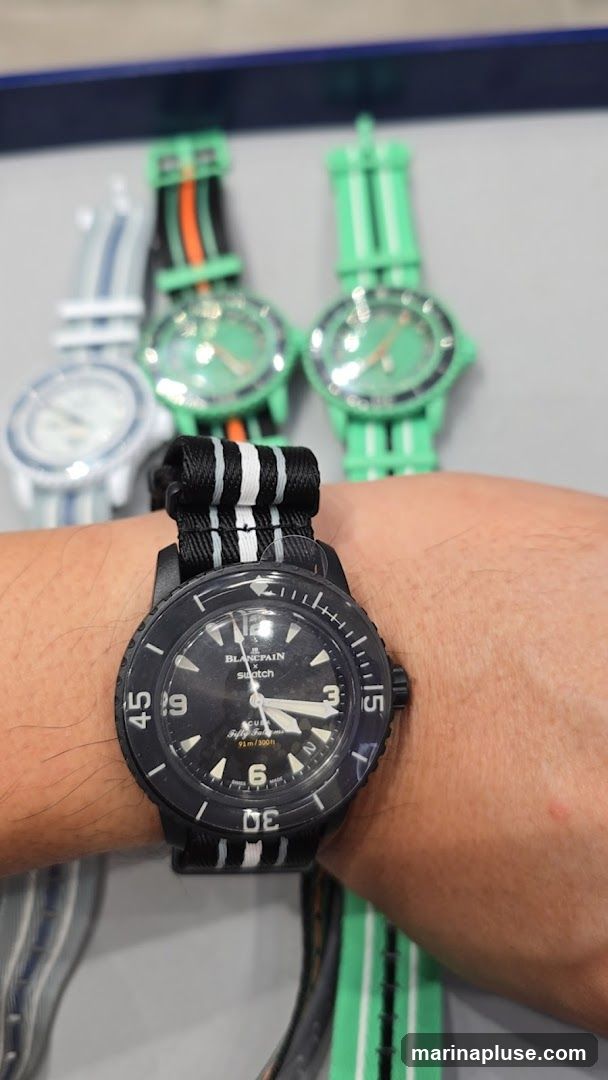 A close-up shot of a person wearing a black Blancpain x Swatch watch on their wrist. The watch has a black dial with white markers and hands, and the bezel is marked with numbers. The strap is black with white stripes. In the background, several other Swatch watches with green straps and green dials are visible.
