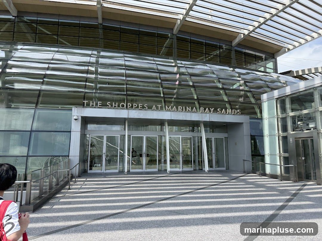 A wide shot of the exterior of The Shoppes at Marina Bay Sands, a modern shopping complex with a glass facade. The name of the establishment is prominently displayed in large silver letters above the entrance. The entrance features multiple glass doors and is flanked by glass walls. To the right of the entrance, there is a glass elevator. The ground in front of the entrance is paved with a speckled material, and diagonal shadows from an overhead structure create a striped pattern. A person with a red backpack is partially visible on the left side of the frame.