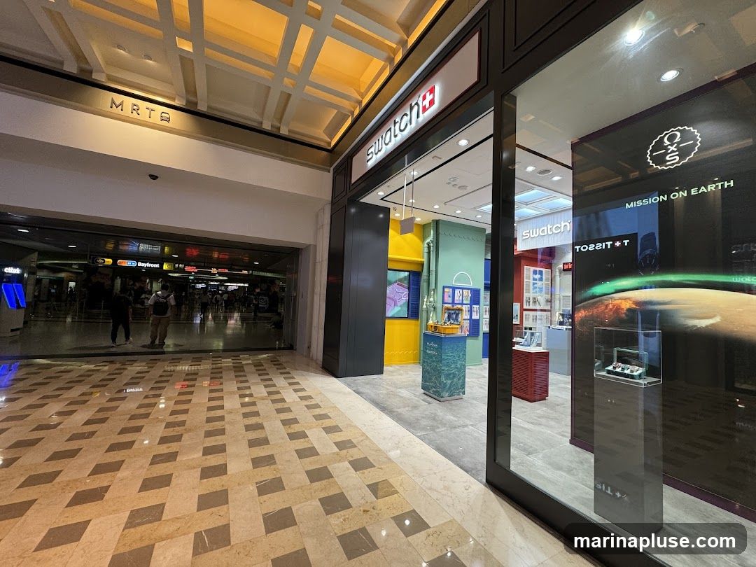 The exterior of a Swatch store is visible, with its distinctive red and white logo above the entrance. The store features large glass windows displaying watches and promotional materials. To the left, a sign indicates "MRT Q". The interior of the store is brightly lit and showcases various watch displays.