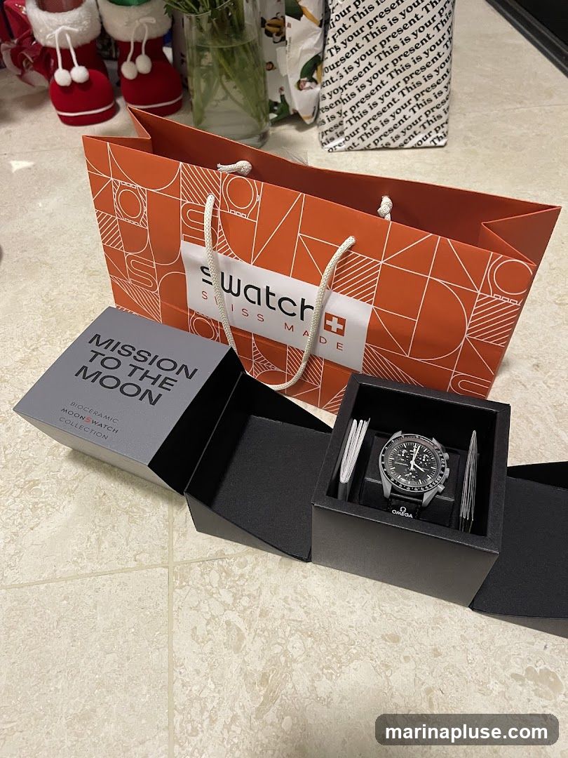 A Swatch "Mission to the Moon" Bioceramic Moonswatch is displayed in its open black presentation box. The watch has a black dial and a silver-toned case and bracelet. Next to the watch are two small booklets. In the background, an orange Swatch shopping bag with a geometric pattern and the Swatch logo is visible. Also in the background are Christmas stockings and wrapped gifts.