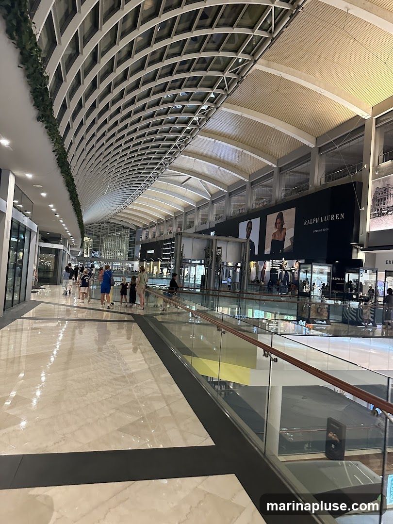A wide, well-lit walkway inside a shopping mall with a distinctive curved ceiling structure. People are seen walking along the corridor, and various shop fronts are visible on either side. A large black banner with "RALPH LAUREN" and "OPENING SOON" is prominently displayed. The floor is made of polished marble tiles.