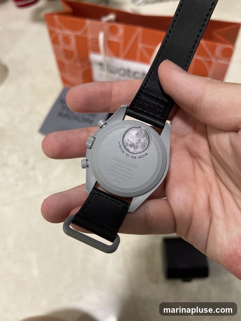 A person is holding the back of a Swatch watch with a black strap. The watch case is light gray and has text engraved on it, including "MISSION TO THE MOON" and "Swatch® SWISS MADE WATER-RESISTANT". In the background, there is an orange shopping bag with the Swatch logo visible.