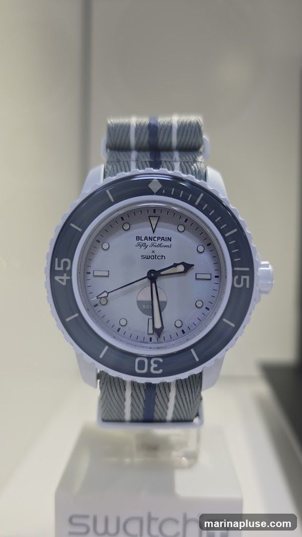 A close-up shot of a white Blancpain Fifty Fathoms x Swatch watch with a grey and blue striped fabric strap. The watch face is white with silver hour markers and hands. The bezel is dark grey with white numerals. The watch is displayed on a white stand with the Swatch logo visible at the bottom.