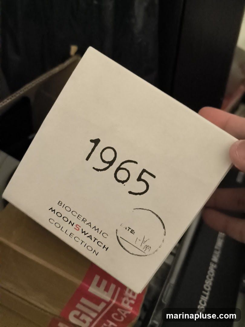 A white box with the number 1965 printed in large black font. Below the number, it reads "BIOCERAMIC MOONSWATCH COLLECTION". There is a circular stamp with the word "DATE:" and a handwritten date. The box is held by a hand and is partially in a cardboard box with red "FRAGILE HANDLE WITH CARE" tape.