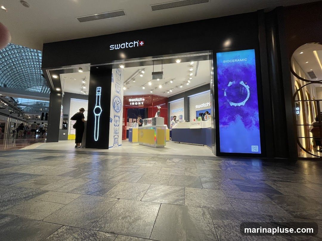 The exterior of a Swatch store with its prominent signage. The store features a large digital display on the right showcasing a watch design with the text "BIOCERAMIC". To the left, the store entrance is visible with a large illuminated outline of a watch on a pillar. Inside, the store is brightly lit, displaying various watches and merchandise. The Swatch logo and "swatch+ IRONY" are clearly visible on the facade.