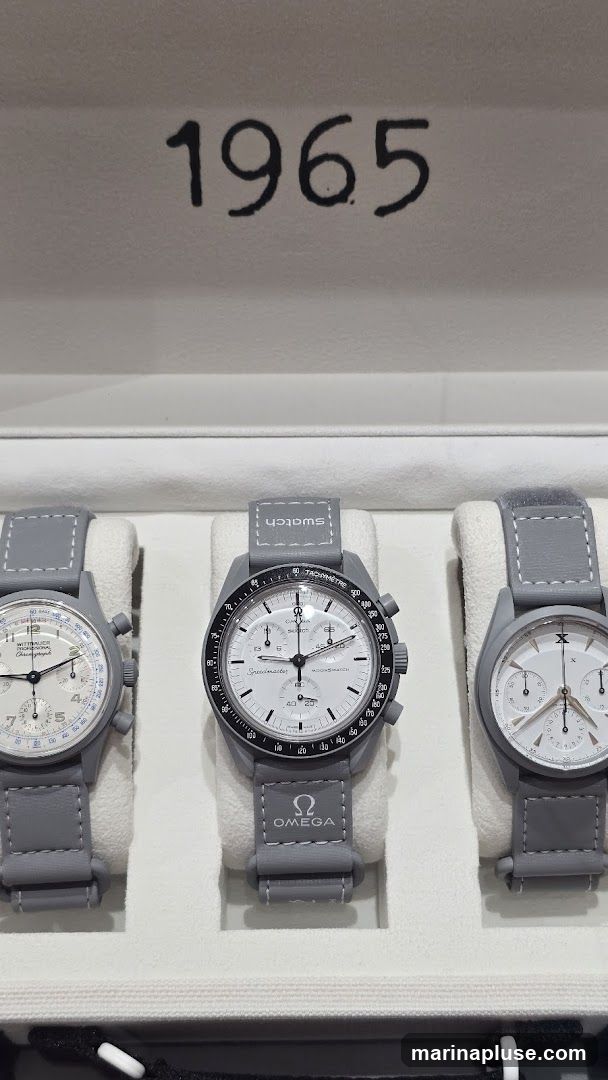 Three grey watches with white faces are displayed in a white box. The watches have grey straps. The middle watch has the OMEGA logo and the word Speedmaster printed on it. The word SWATCH is printed on the strap of the middle watch. The number 1965 is printed in black on the inside lid of the box.