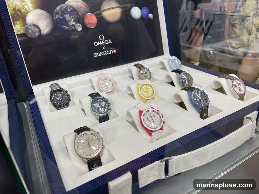 A display case filled with various Omega x Swatch watches, each presented on a white stand. The background features a graphic of planets and the Omega x Swatch logo. The watches are in different colors, including black, green, pink, yellow, blue, red, white, and beige.