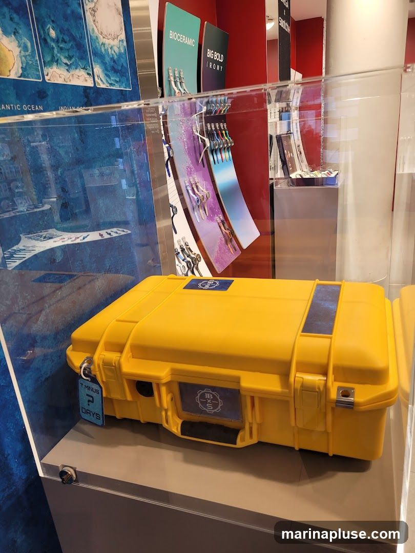 A bright yellow, rugged protective case is displayed inside a glass enclosure. The case has two blue and silver emblems on it, one on the lid and one on the front. A tag attached to the side of the case reads "T MINUS ? DAYS". In the background, there are displays of watches and artwork, including what appears to be a map of the ocean.