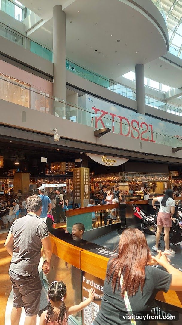A bustling indoor food court with multiple dining options and shoppers. A sign for "KAUAN MASTERS" is visible above one of the eateries. In the foreground, a family is interacting with a display. Another large sign reads "KIDS21".