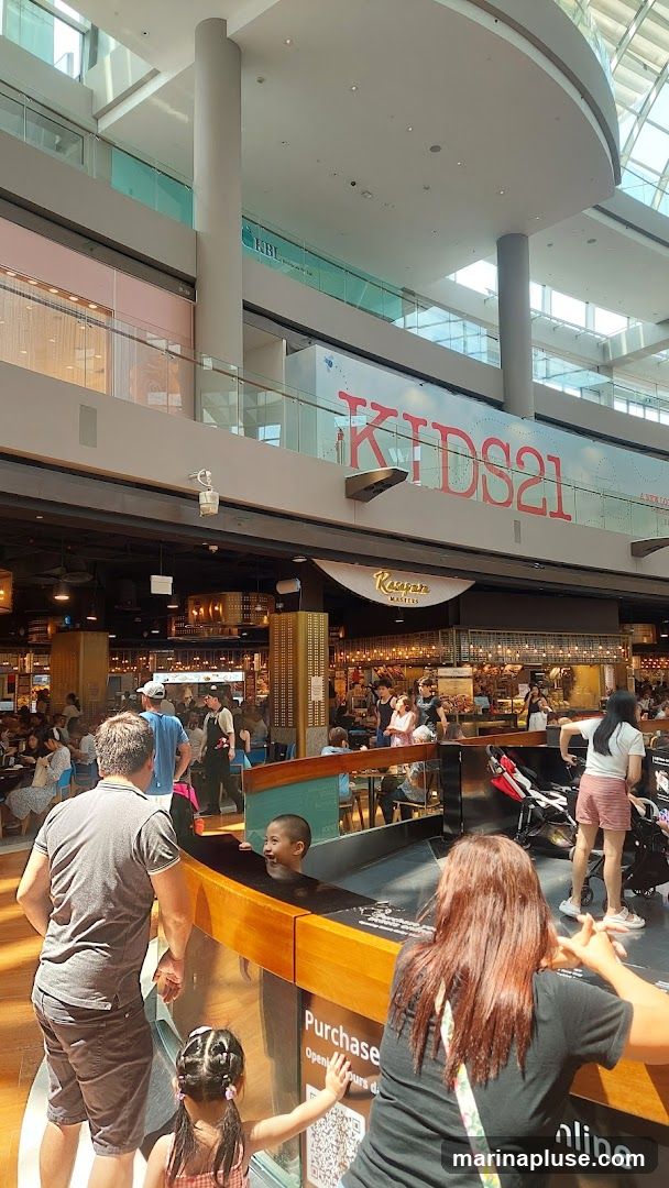 A bustling indoor food court with multiple dining options and shoppers. A sign for "KAUAN MASTERS" is visible above one of the eateries. In the foreground, a family is interacting with a display. Another large sign reads "KIDS21".