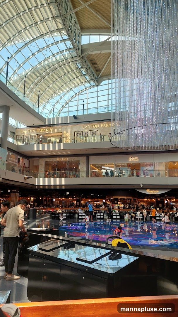 An interior view of a large, modern shopping mall with a high, arched glass ceiling. Multiple levels of shops are visible, including