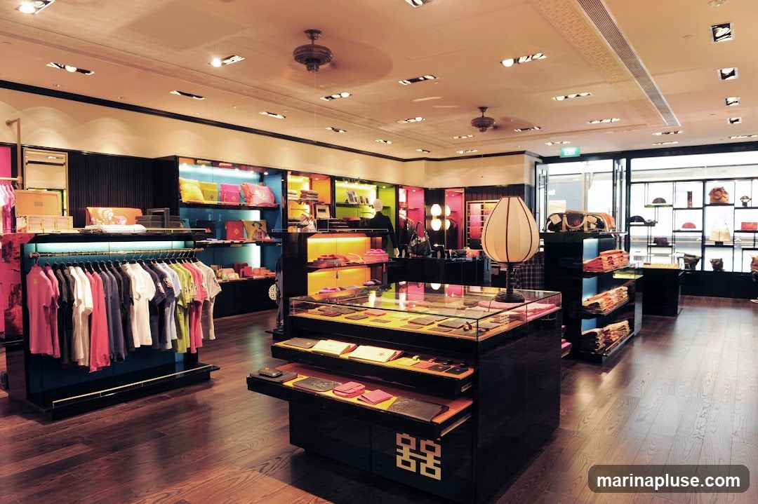 A well-lit, upscale retail store interior featuring clothing racks with colorful t-shirts, display cases with wallets and accessories, and shelves stocked with bags and other merchandise. The store has a modern design with dark wood flooring, black display units, and brightly colored shelving. A large, decorative lamp stands prominently in the foreground.