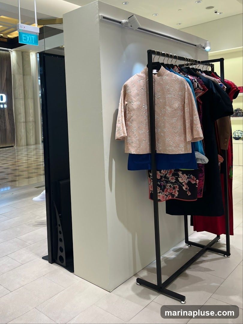 A clothing rack displays several garments, including a light pink patterned jacket, a blue garment, and a skirt with a floral pattern. The rack is positioned in front of a cream-colored partition. In the background, an illuminated 'EXIT' sign is visible, along with a glimpse of a shopping mall interior.