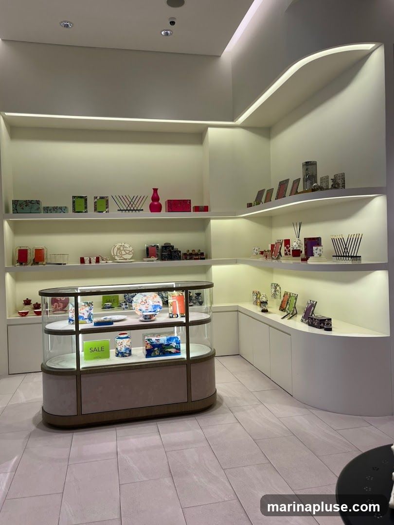 A well-lit store interior with shelves and a display case filled with various decorative items. The shelves display picture frames, vases, boxes, and other small ornaments. The glass display case showcases plates, vases, and boxes, with a prominent green 'SALE' sign on one shelf. The flooring is tiled.