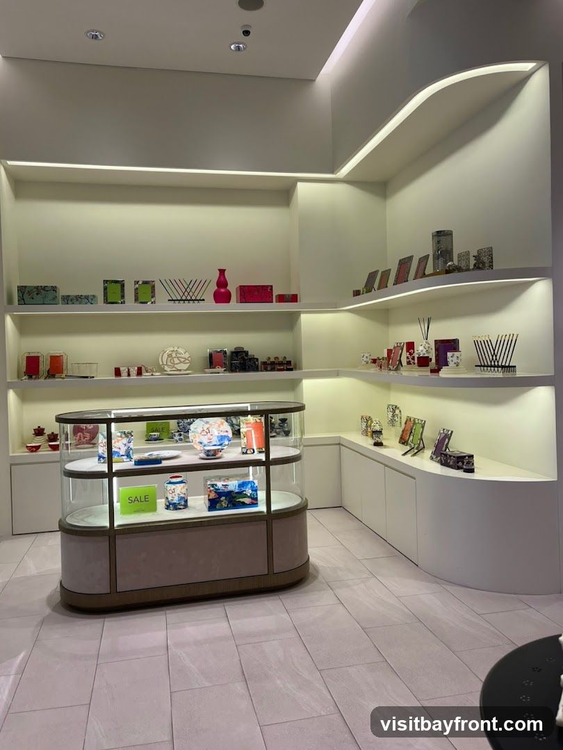 A well-lit store interior with shelves and a display case filled with various decorative items. The shelves display picture frames, vases, boxes, and other small ornaments. The glass display case showcases plates, vases, and boxes, with a prominent green 'SALE' sign on one shelf. The flooring is tiled.