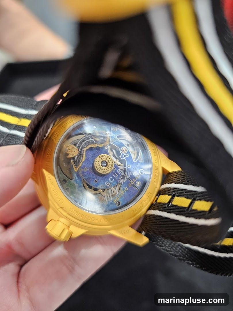 A close-up shot of the back of a yellow watch case. The caseback is transparent, revealing the intricate inner workings of the watch. Text around the edge of the caseback reads "LICENCE TO EXPLORE OCEAN BREATH MERSE YOURSELF 2006/06 PASSION FOR DIVING". The watch has a black and yellow striped fabric strap.
