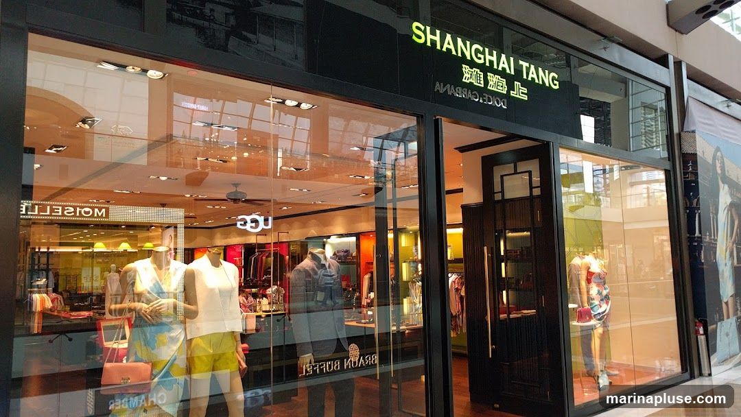 The exterior of a Shanghai Tang store, featuring a large glass window displaying mannequins dressed in clothing and accessories. The store's name is prominently displayed in green lettering above the entrance. Reflections of the interior and surrounding mall are visible on the glass.