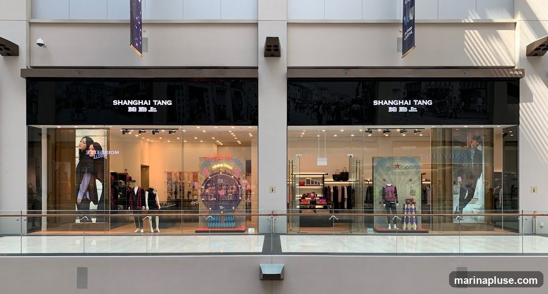 A wide shot of the Shanghai Tang storefront in a mall. The storefront features large glass windows displaying mannequins in fashionable clothing and various merchandise. Above the windows, the Shanghai Tang logo is prominently displayed in white lettering on a black background, accompanied by Chinese characters. The interior of the store is visible, showing racks of clothing and display shelves.