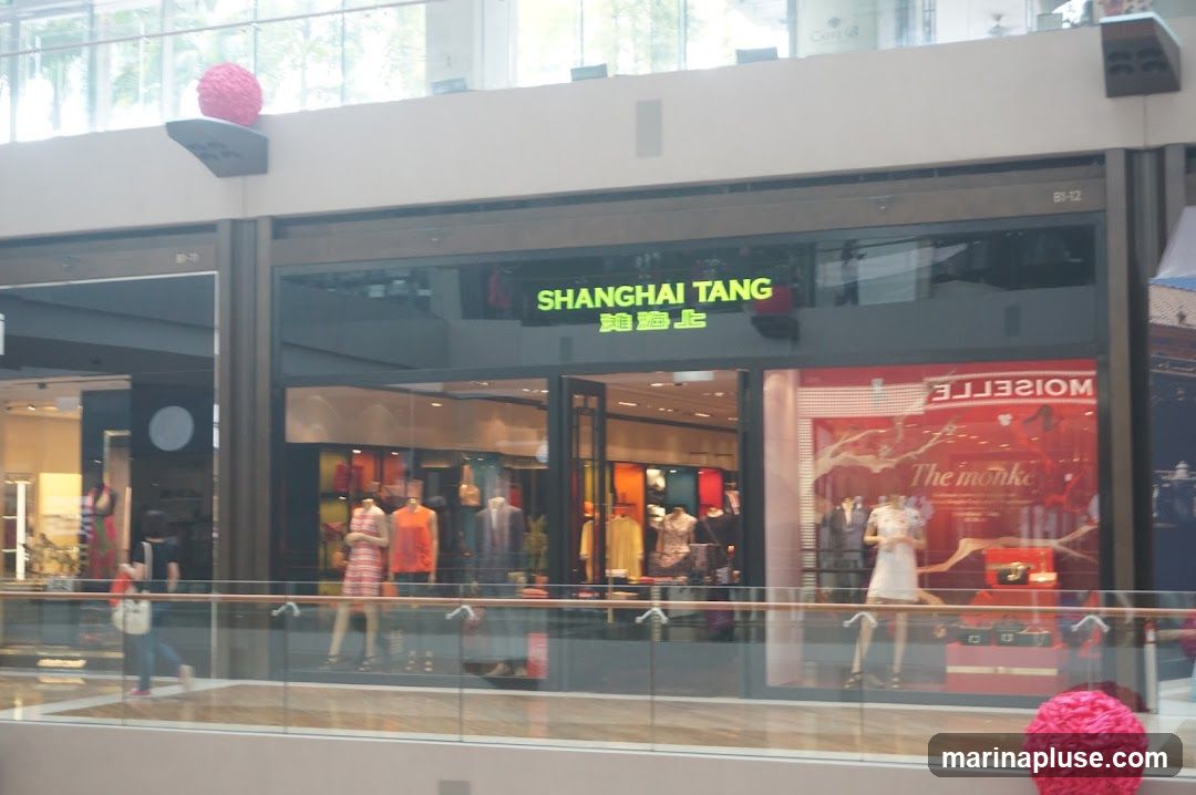The exterior of a Shanghai Tang store is visible, with the store name illuminated in green neon above the entrance. The storefront features large glass windows displaying mannequins dressed in various clothing items. To the right, a window display has a red background with text that reads "The monkey" and other mirrored text. Pink decorative spheres are visible on ledges above and in front of the store.