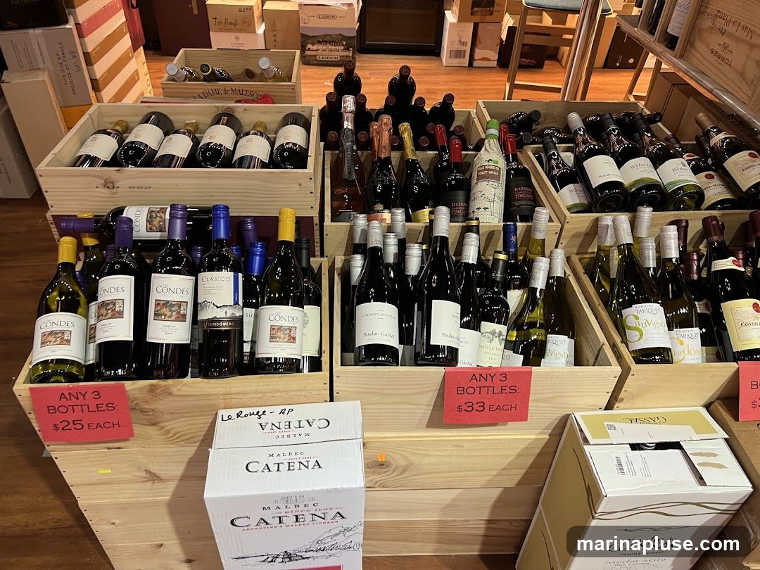 A display of various bottles of wine in wooden crates. Some bottles are arranged in rows, while others are stacked. There are promotional signs indicating prices for multiple bottles. The background shows more shelves and boxes, suggesting a retail environment.
